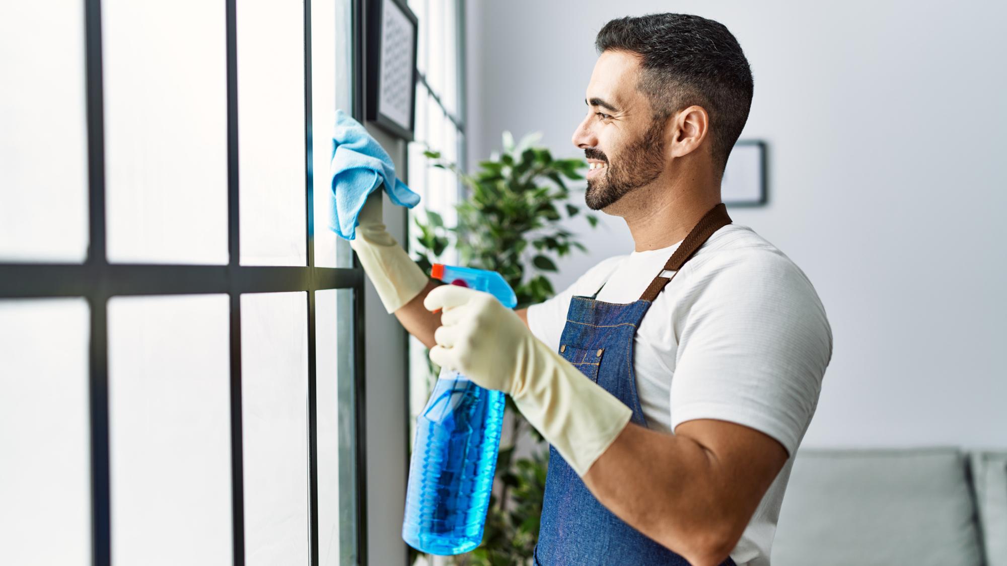 A man wearing gloves and an apron is smiling while cleaning a window with a cloth and a spray bottle in a bright, modern room.